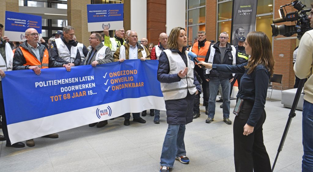 Protesters hold a large blue banner with Dutch text; a female reporter interviews a man in a white hi-vis vest as a news crew films.