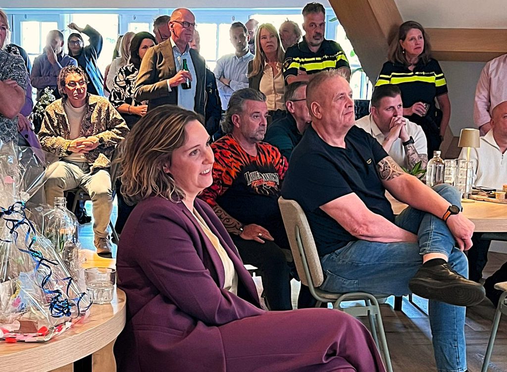 Group of people at an indoor gathering watching something off-camera; foreground features a woman in a purple suit smiling. Earlier, a man in a black shirt with tattoos sits at a table with drinks and gifts nearby.