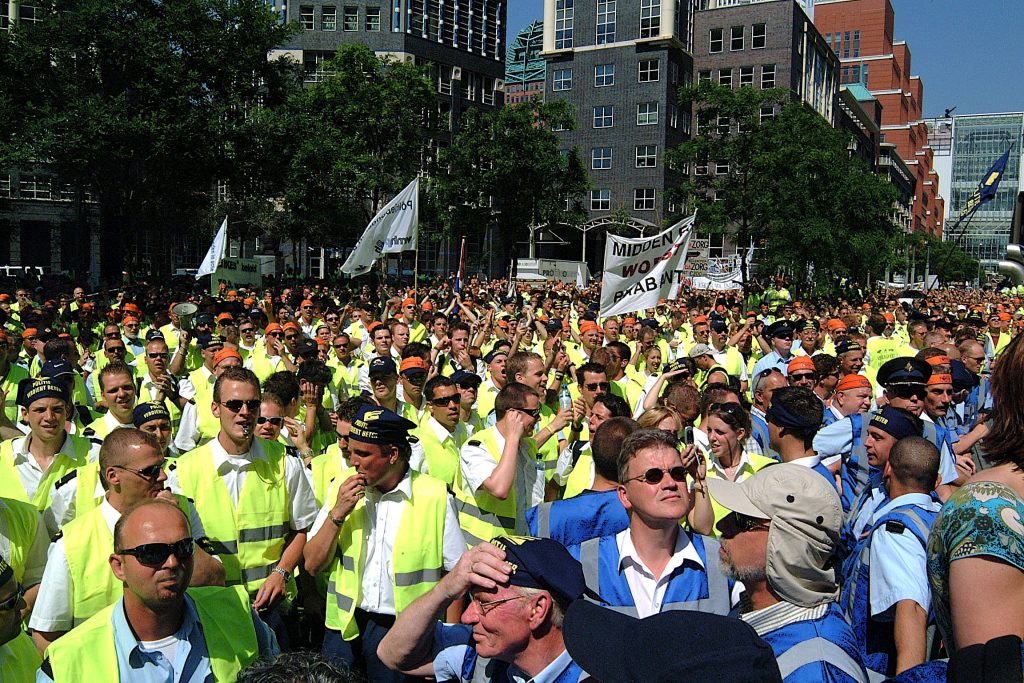 Large crowd of people wearing high-visibility vests in a street protest, holding banners and flags with city buildings in the background.