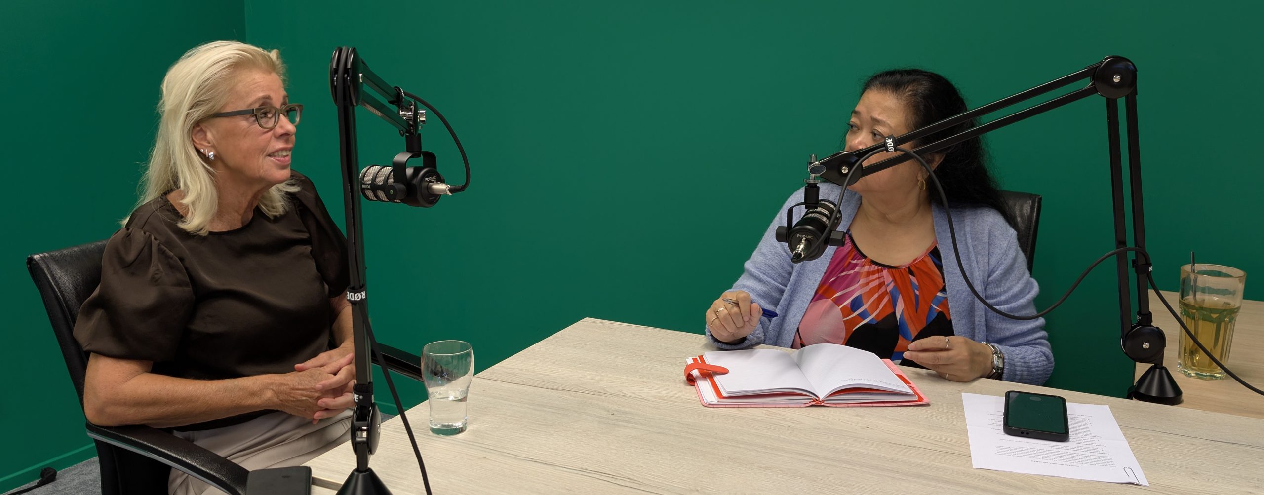 Two women sit at a table in a recording studio with boom microphones, talking to each other. The older woman on the left speaks while the younger woman on the right takes notes in a red-edged notebook.