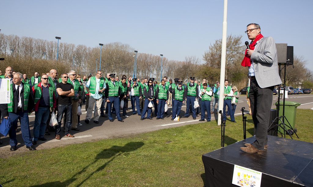 Speaker on a stage addressing a crowd of volunteers wearing green reflective vests outdoors at an event.