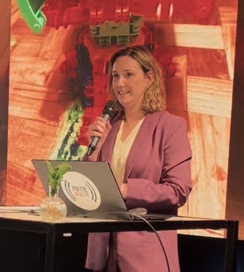 Woman in a pink blazer speaks into a microphone at a podium on a stage, with a laptop and a small plant in front of her.