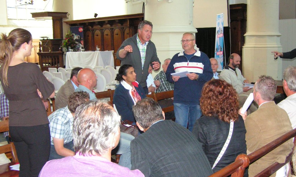Man standing and pointing while speaking to a seated audience in a hall with pews and a stage in the background.