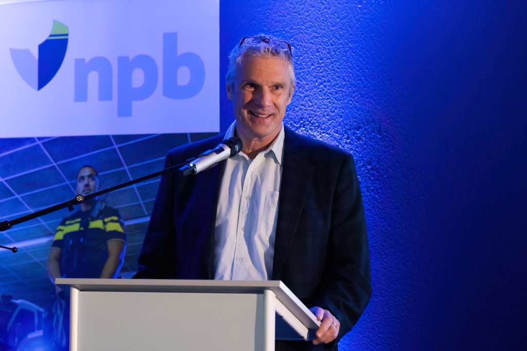 Man in a suit speaking at a podium with a microphone, blue stage lighting and a banner in the background.