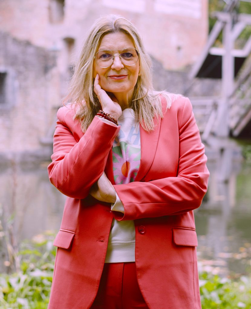 Portrait of a smiling woman with blond hair and glasses, wearing a pink blazer, standing by a canal outdoors.
