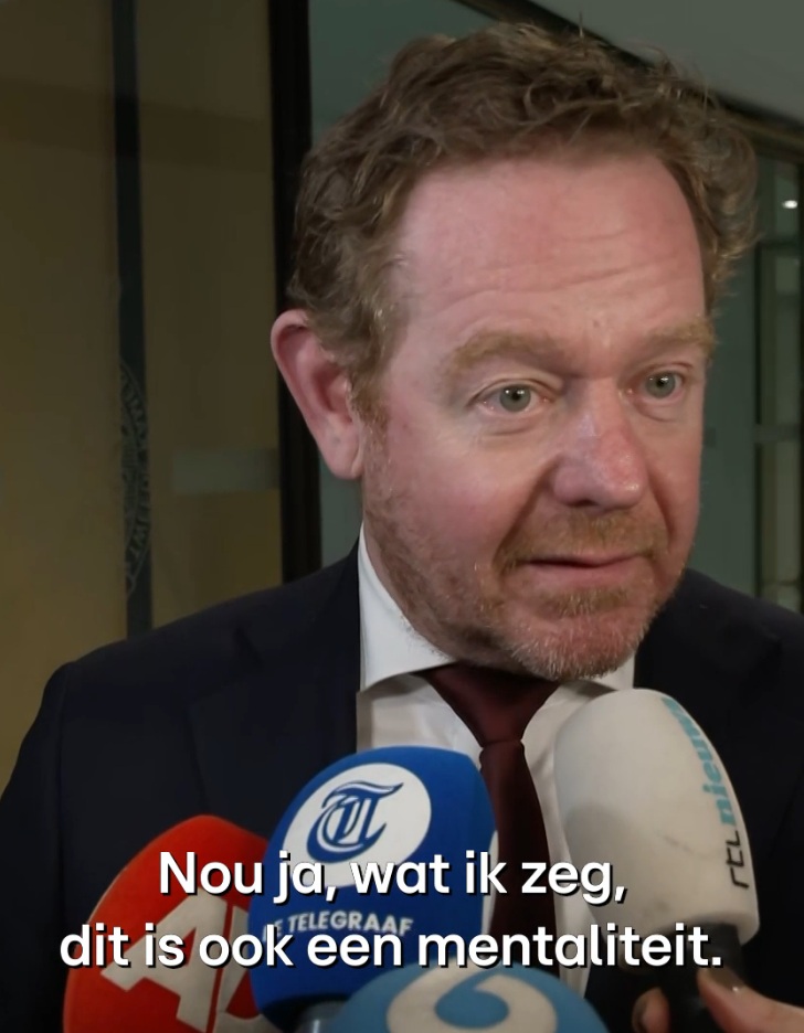 Man in a suit being interviewed by several reporters, microphones in front; Dutch subtitle about mentality visible.