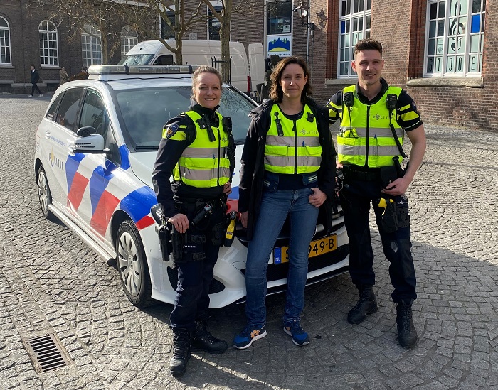 Three Dutch police officers in high-visibility vests pose beside a marked police car on a cobblestone square.