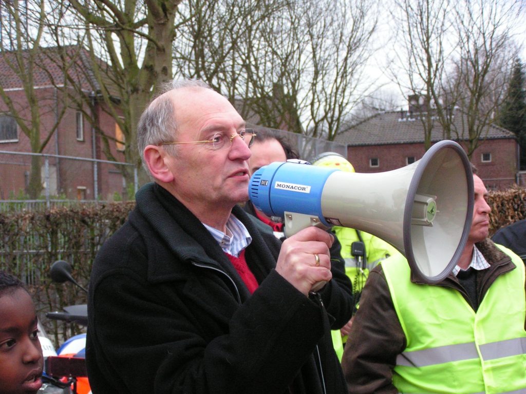 Man with glasses speaks into a blue megaphone at an outdoor event, with others in high-visibility vests and leafless trees in the background.