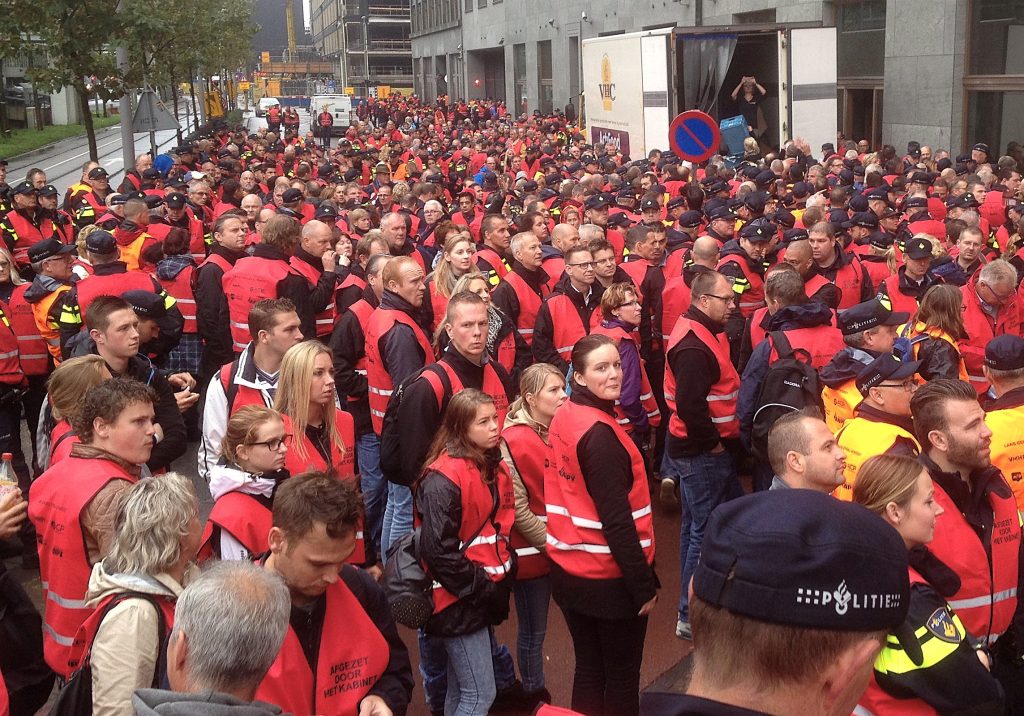 Large crowd of people wearing red safety vests fills a city street, gathered for a protest or event near a truck and tall buildings.