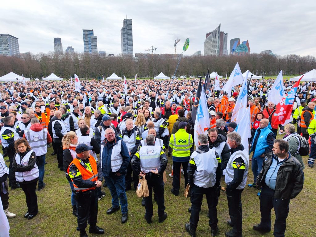Large crowd of people in high-visibility jackets at an outdoor protest or rally, many holding flags on a park lawn with city buildings in the background