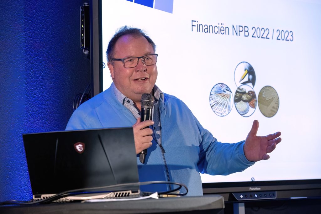 Presenter in a blue jacket speaks into a microphone in front of a slide labeled 'Financiën NPB 2022/2023' with coin graphics.