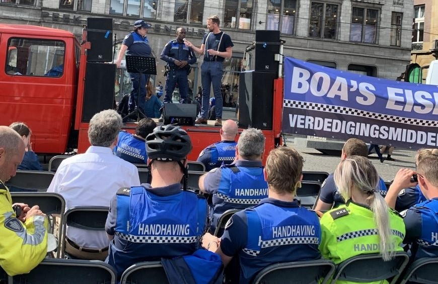 Uniformed officers on a red flatbed stage speak to a seated outdoor crowd, with a blue banner and speakers behind them.