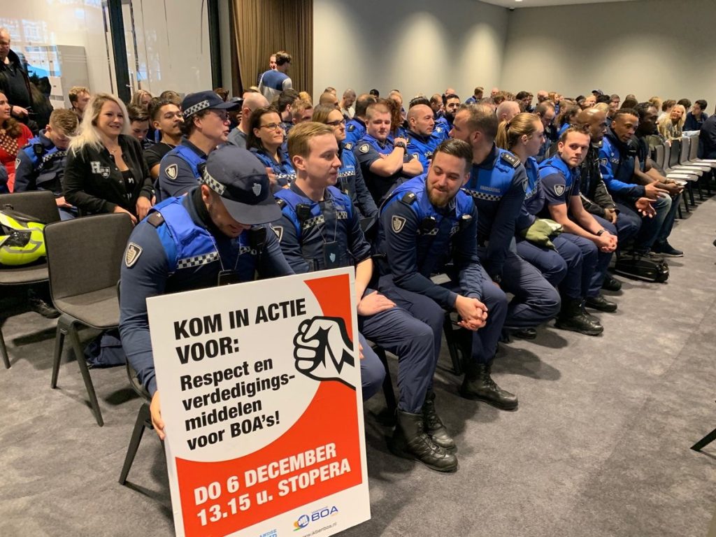 Group of uniformed security officers seated in a conference room; one holds a protest sign about respect and protective gear for BOA officers, with event details on the poster.