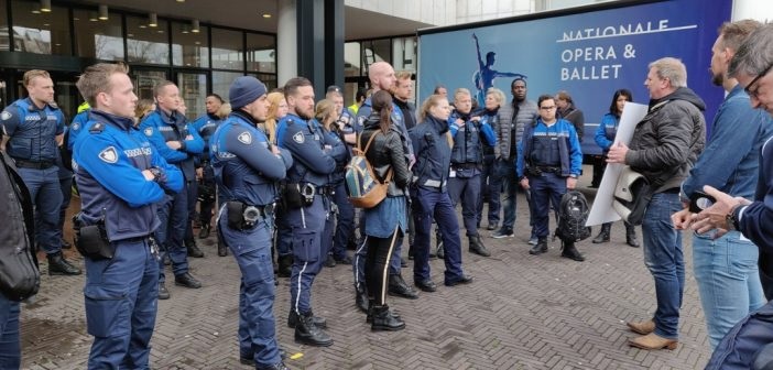 Uniformed officers stand in a semicircle outside a building, listening to a speaker with rolled posters near a National Opera & Ballet banner.