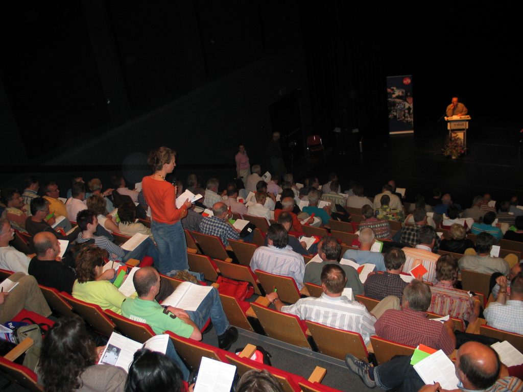 Audience seated in a dark auditorium, many holding papers, facing a stage with a speaker at a podium on the right.