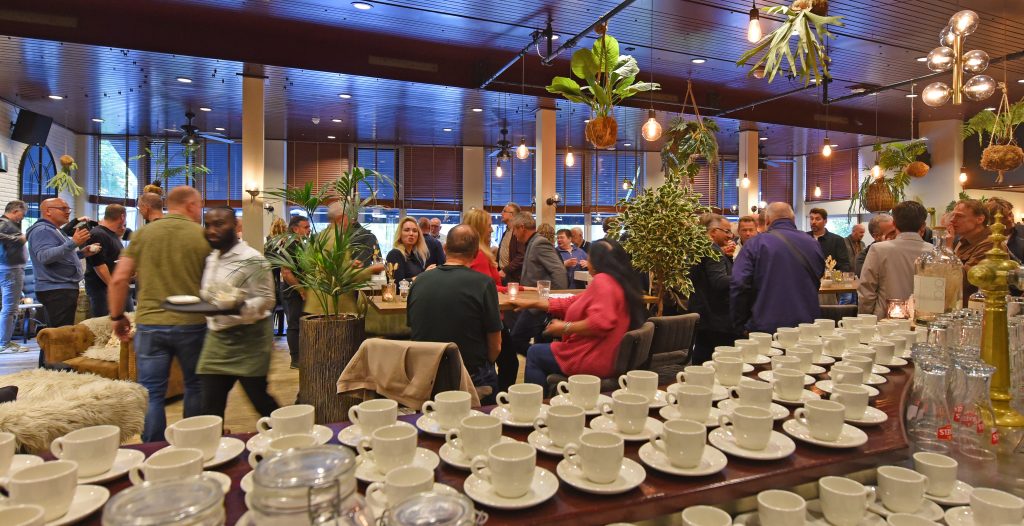 Busy social event in a modern cafe; people mingle around a long table of white coffee cups and plants under warm hanging lights.