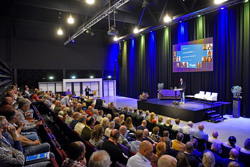 Conference hall with stage, speaker at a podium, and a large screen; audience seated facing the stage.