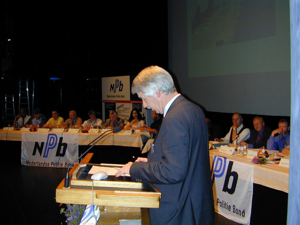 Speaker at a wooden podium addressing a long panel of people on stage, with NPB banners reading 'Nederlandse Politie Bond' in the background.