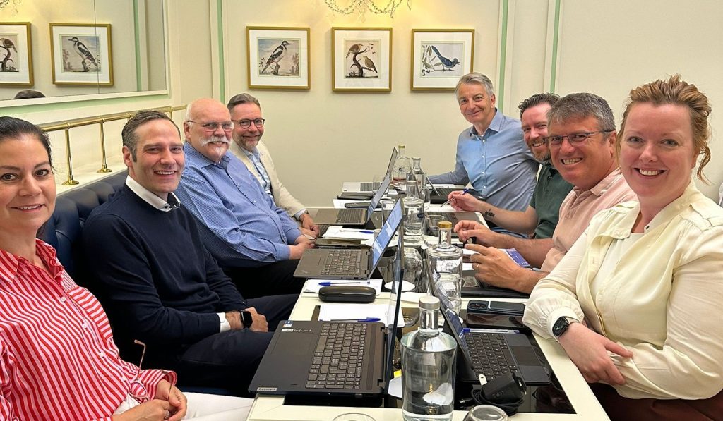 Eight people in a business meeting around a long table with laptops and water bottles in a bright room with framed bird artwork on the wall.