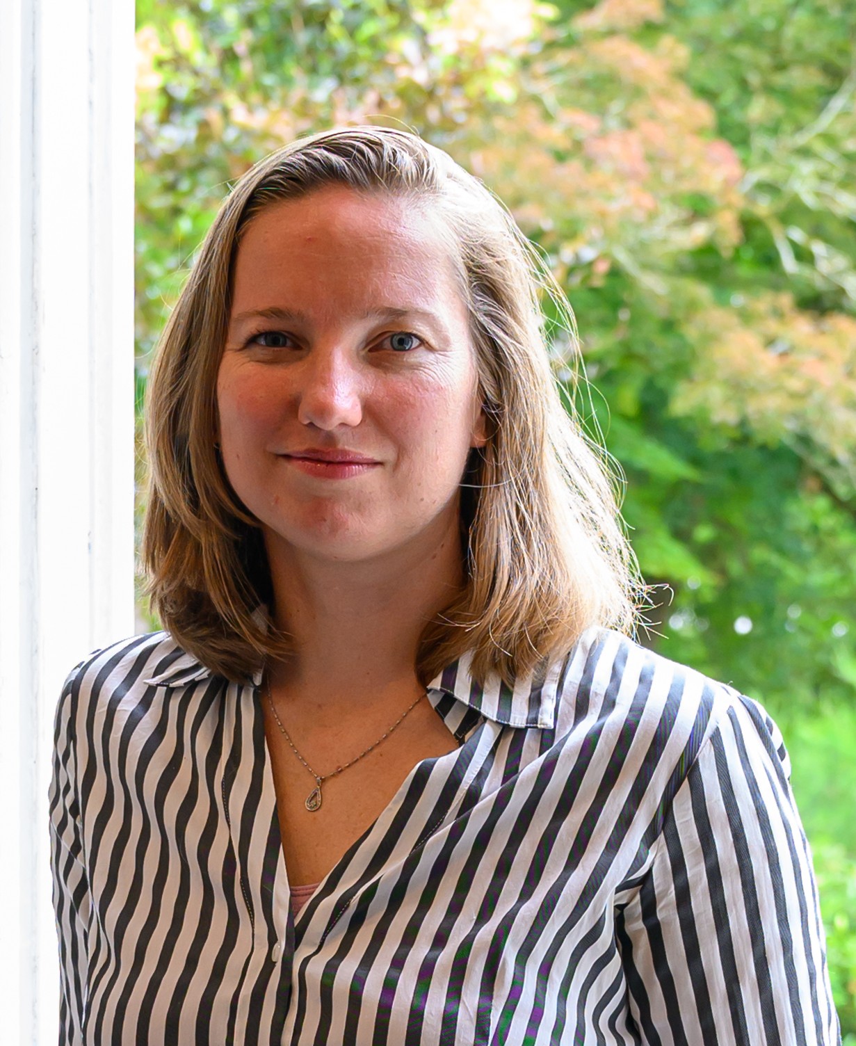 Woman with shoulder-length blond hair wearing a striped black-and-white blouse, standing by a window with green foliage outside.