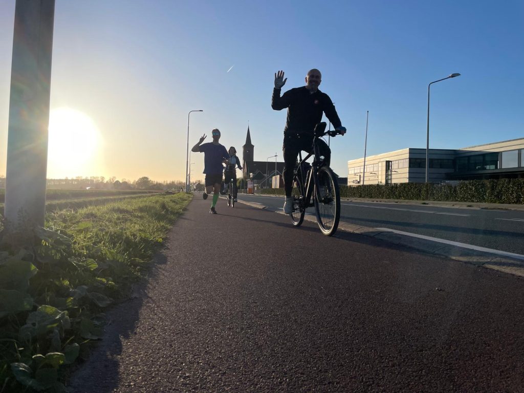 Three people biking along a sunlit sidewalk on a clear day, the front rider waving at the camera with a church spire in the background.