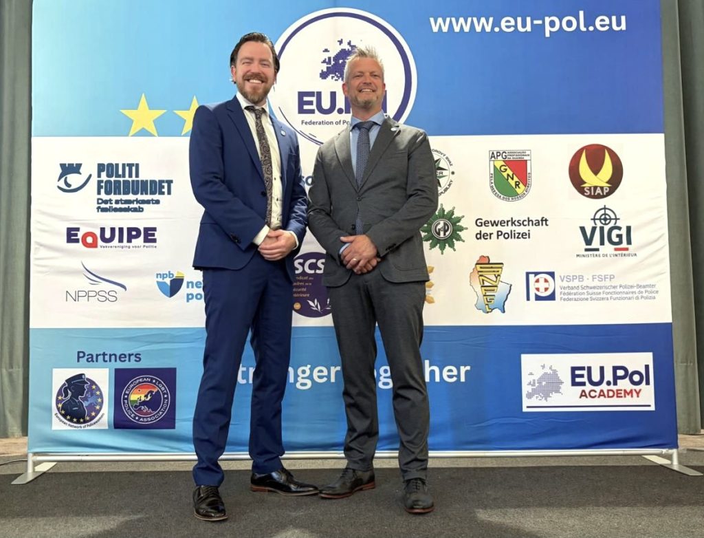 Two men in suits pose and smile in front of a blue EU-Pol event banner filled with logos and stars.