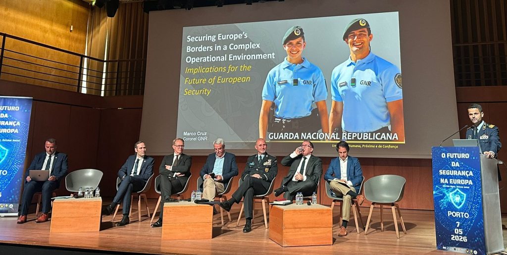 Panel discussion on stage with seven men in business attire; a large screen shows two uniformed guards and a security conference title in Portuguese/English text.