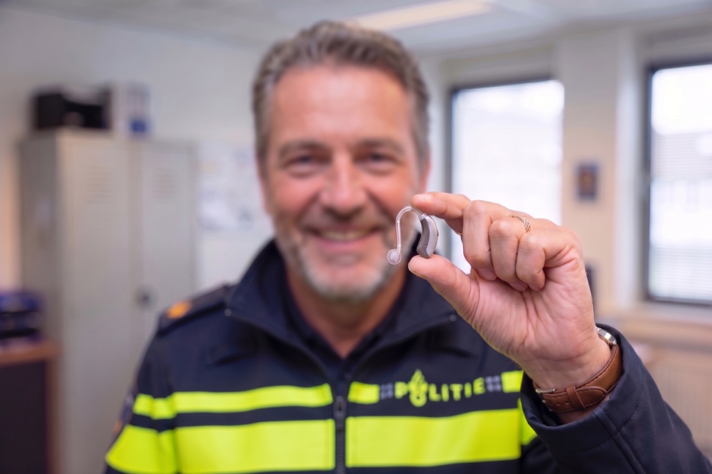 Smiling man in a high-visibility uniform showing a clear behind-the-ear hearing aid to the camera.
