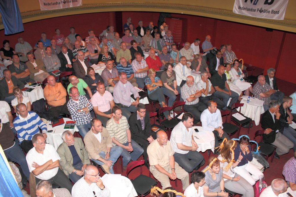 Large audience seated in an auditorium watching an event, with tables and papers in front of some attendees and banners overhead.