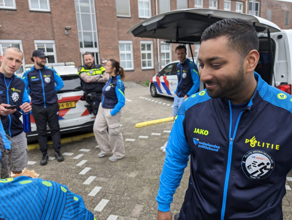 Group of uniformed emergency responders standing outside a brick apartment building near a police van in the background.