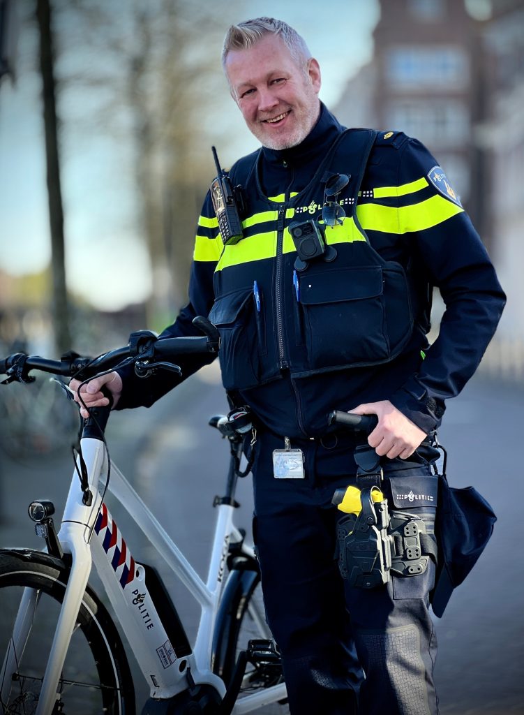 Police officer in a high-visibility jacket standing with a white patrol bike, smiling at the camera outdoors.