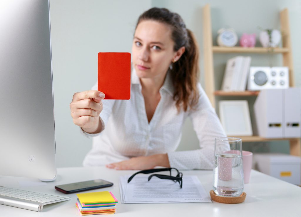Woman at a desk holding up a red card toward a computer monitor in an office setting.