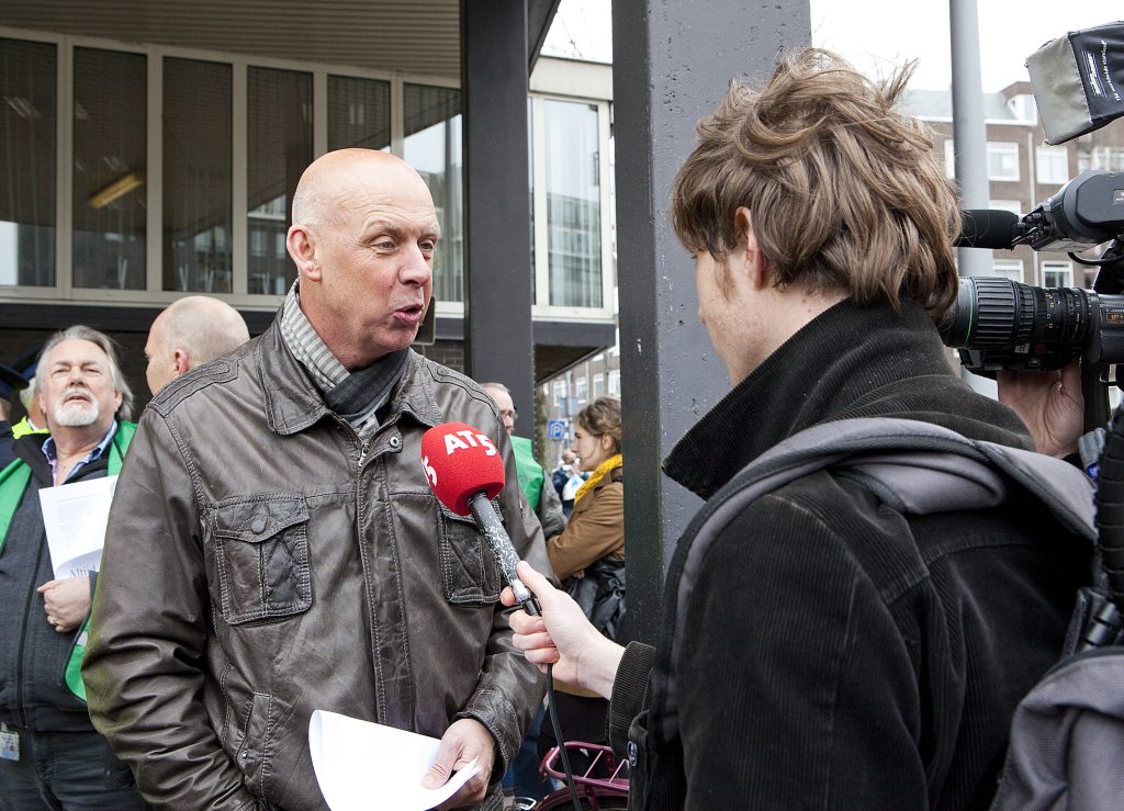 Man with a brown leather jacket being interviewed outdoors, holding papers, as a reporter with a red mic and a nearby camera crew record the scene.