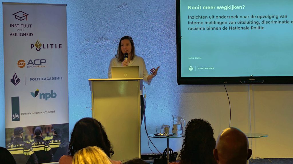 Female presenter at a podium giving a talk, with banners showing police logos on the left and a projection screen on the right.