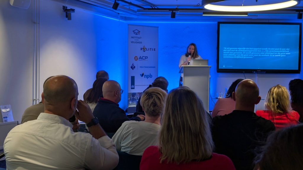 Female presenter at a lectern giving a talk in a blue-lit conference room, audience seated facing the stage with a projection screen to the right and a banner with logos nearby.