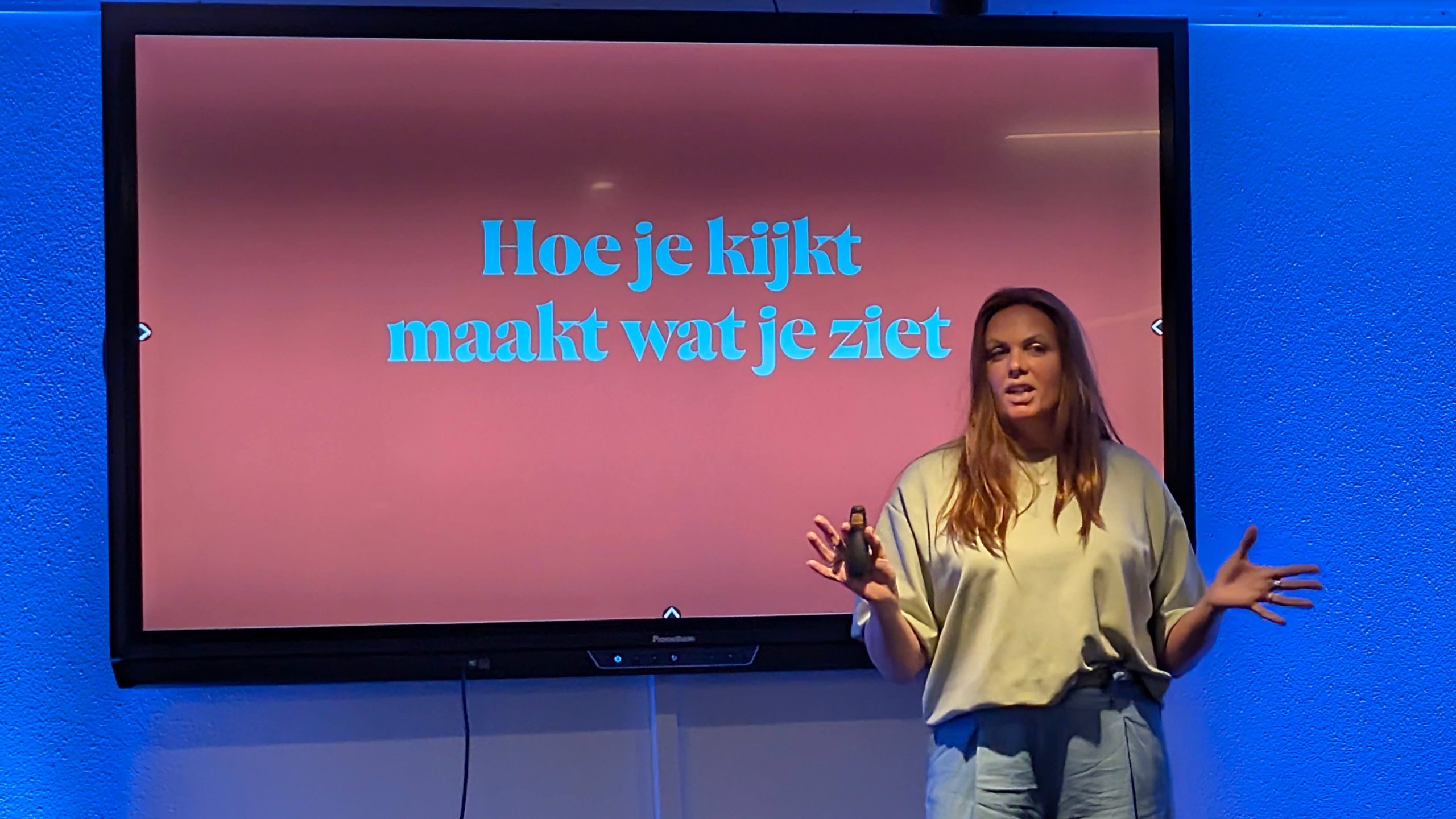 Woman giving a talk in front of a monitor displaying a pink slide with the Dutch phrase 'Hoe je kijkt maakt wat je ziet'.