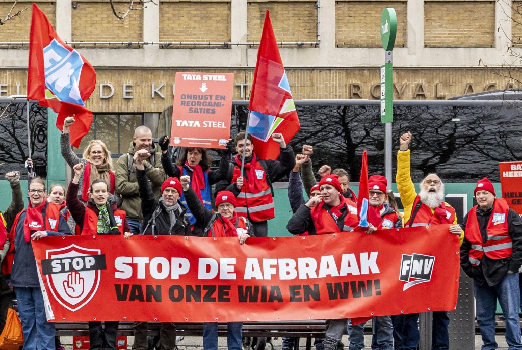 Group of protesters on a city street holding a large red banner that reads 'STOP DE AFBRAAK' with red flags and FNV logos visible.