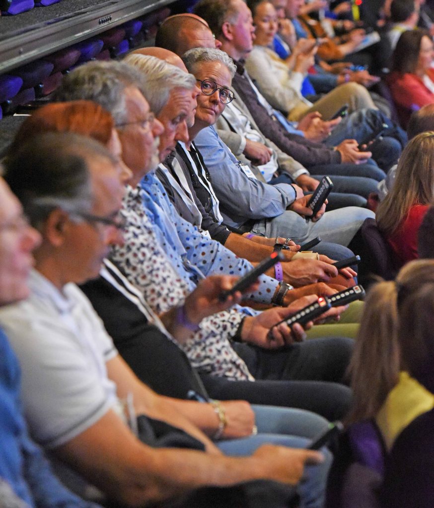 Audience seated in a theater or conference venue, many people looking toward the stage; several hold smartphones or remote-like devices in their hands.