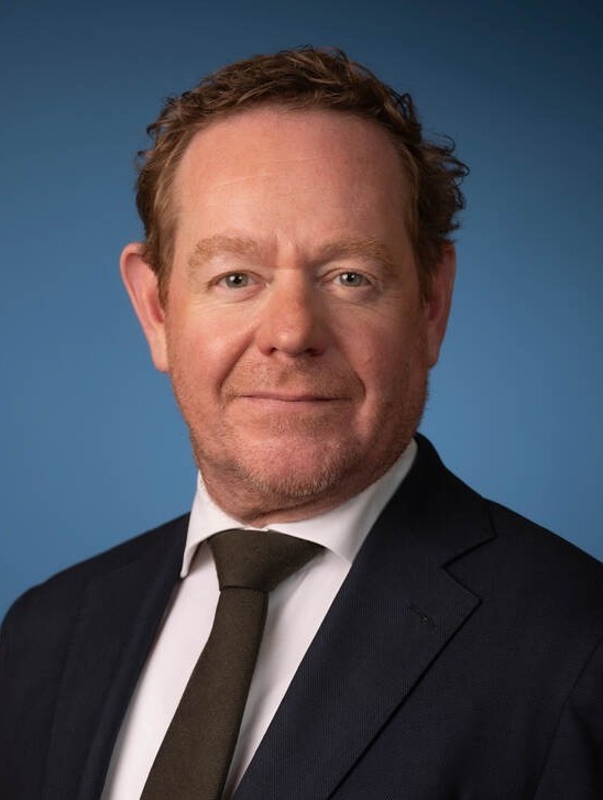 Professional headshot of a middle-aged man in a dark suit and olive-brown tie, against a blue backdrop.