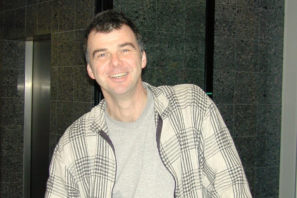 Smiling man in a gray shirt and plaid jacket standing in a building lobby with dark tile walls behind him