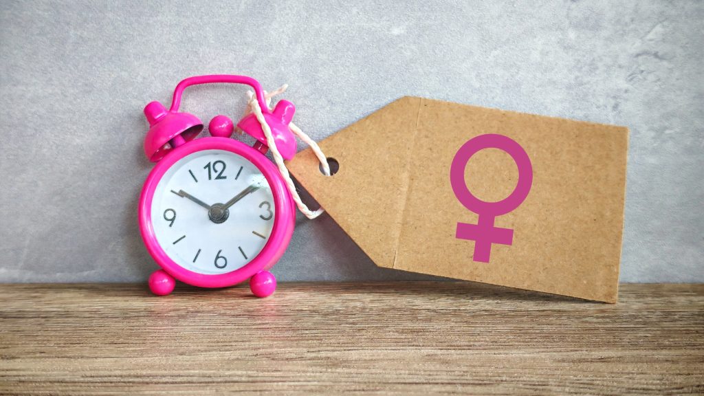 Pink alarm clock beside a cardboard tag bearing a purple female gender symbol on a wooden surface.