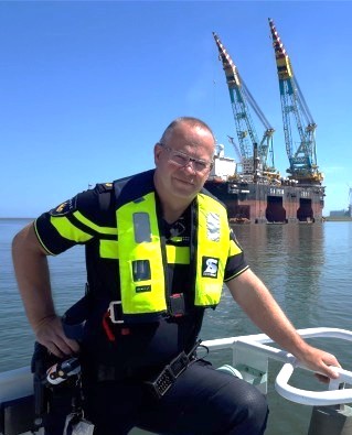 Man wearing a bright yellow life vest on a small boat, with an offshore oil rig in the blue sea behind him.