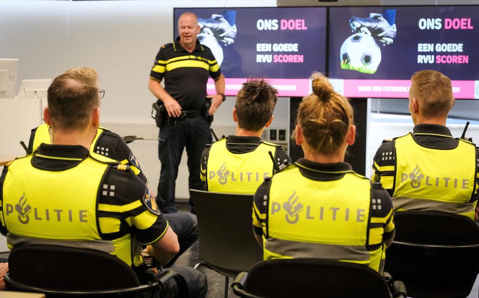 Police officers in bright yellow vests sit in a row, facing a presenter in a briefing room with dual screens behind him, displaying 'ONS DOEL'.