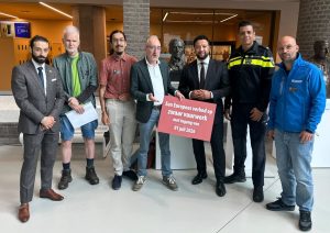 Eight men stand in a lobby; one holds a red sign about a European heavy-fireworks ban starting July 1, 2026.