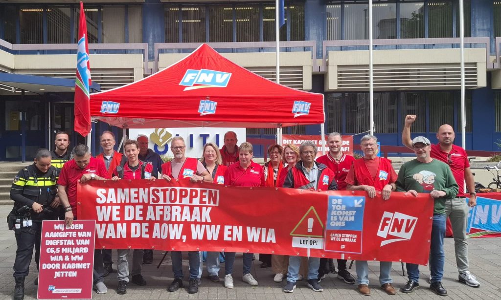 Group of people in red shirts and vests hold a large red protest banner with FNV logos outside a building.
