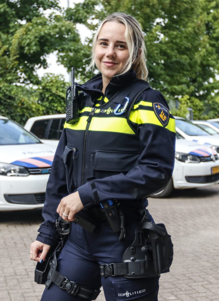 Smiling female police officer in a navy uniform with neon yellow stripes, radio on her shoulder and a duty belt, standing outdoors with police cars in the background.