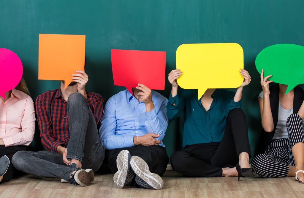 Group of people sitting against a green wall, each holding large colorful speech bubbles that cover their faces.