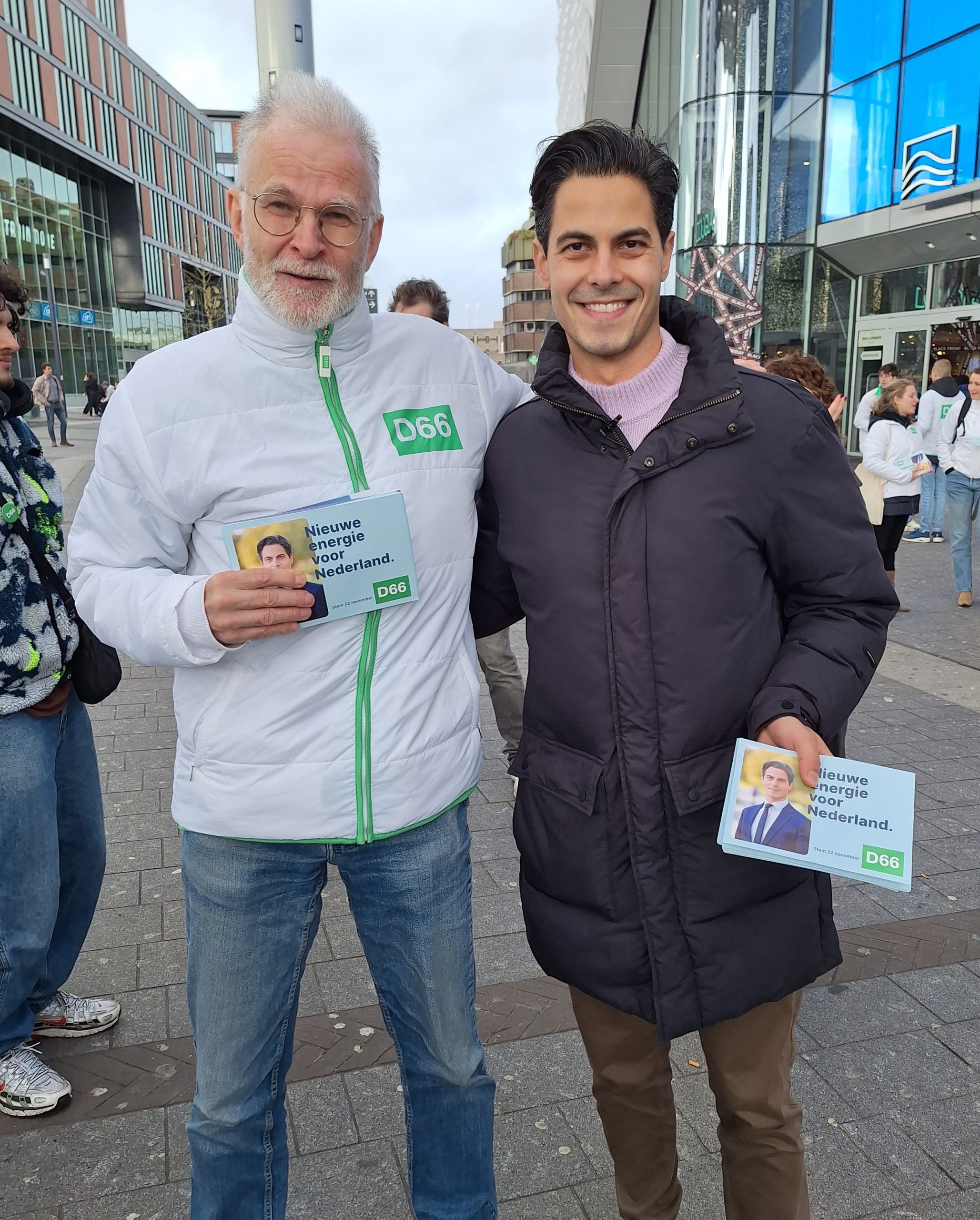 Two men pose together outdoors, campaigners with pamphlets reading 'Nieuwe energie voor Nederland' for D66, in a city plaza.