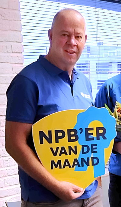 Man in a blue polo shirt holding a yellow and blue award sign reading 'NPB'er van de maand'.