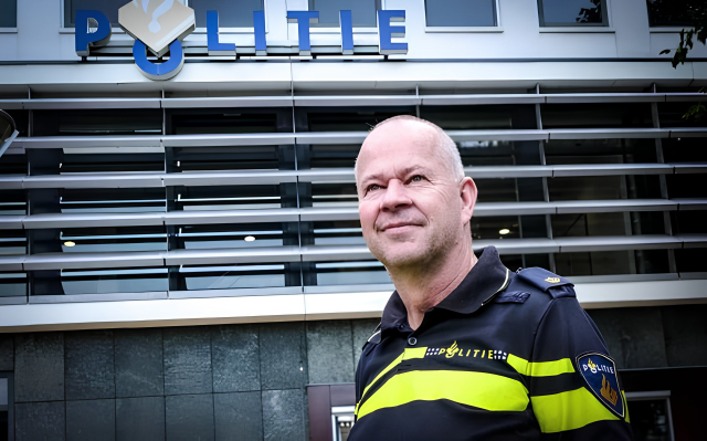 Dutch police officer in uniform stands outside a police station with a blue 'Politie' sign in the background.
